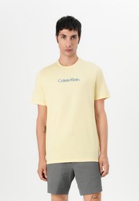 Camiseta de algodón amarilla con cuello redondo, que presenta un logo azul de "Calvin Klein". El modelo lleva pantalones cortos de textura gris.