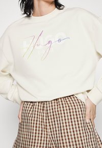Femme portant un sweat-shirt crème avec le texte coloré « Hugo » et « 1983 » en dessous, associé à un pantalon à carreaux marron et beige.