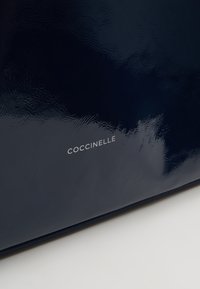 Gros plan d’un matériau bleu marine brillant avec le nom de la marque « COCCINELLE » imprimé en petites lettres blanches.