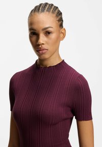 Fioletowa prążkowana bluzka z krótkimi rękawami, z falbaną przy dekolcie i logo na rękawie, wykonana z gładkiej, teksturowanej tkaniny.