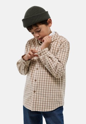 Une chemise à carreaux marron et blanc avec un col et des manches longues, assortie à un jean foncé et un bonnet en tricot vert.