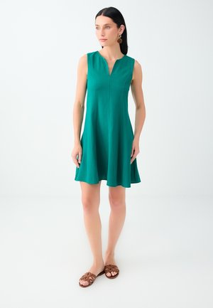 Vrouw staand met een mouwloze groene jurk met een inkeping in de halslijn en bruine platte sandalen op een effen witte achtergrond.