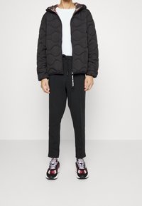 Černá prošívaná zateplená bunda s kapucí, nošená přes bílou tričko. Černé tepláky a tenisky s červenými, bílými a černými akcenty dotvářejí celkový vzhled.