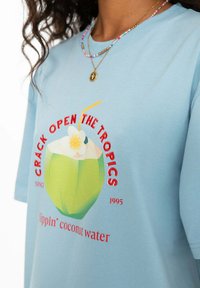 Světle modré bavlněné tričko s grafikou zeleného kokosu se slámkou a květinou, a červeným textem "OTVĚŘTE TROPY."