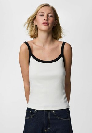 Õlgadeni blond juustega naine, kes kannab valget, musta äärisega varrukatuta t-särki ja tumedaid siniseid teksapükse, seisab ühevärvse tausta ees.