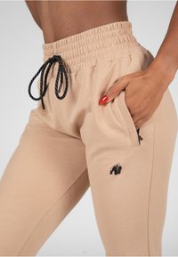 Beige joggingbroek gemaakt van zachte stof met een elastische tailleband, zwarte trekkoord en zijzakken. Beschikt over een klein zwart gorilla logo.