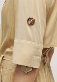 Chemise beige à manches courtes avec un bouton en écaille de tortue qui maintient une manche retroussée au-dessus du coude.