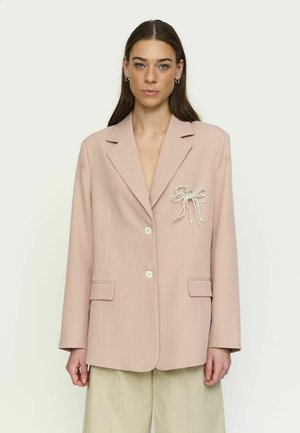 Vrouw met lang bruin haar draagt een beige blazer met parelstrikbroche en bijpassende wijde broekspijpen tegen een effen achtergrond.