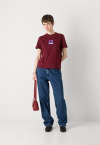 Burgunderfarget bomull T-skjorte med korte ermer, med en lilla CK-logo; kombinert med vide blå denimjeans og en rød håndveske.