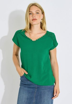 Mujer con cabello rubio que lleva un top verde de manga corta con detalles de encaje y una falda vaquera azul, posando con una mano en el bolsillo.