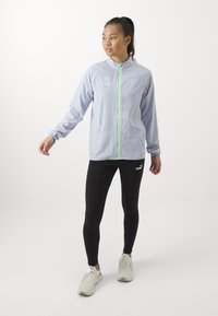 Lyseblå lynlåsejakke med grønne lynlåse, der har sidelommer og logo-detaljer; parret med sorte leggings og hvide træningssko.