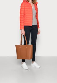 Brun læder tote taske med to lange håndtag, rektangulær form, glat tekstur og minimalistisk design, suppleret af en orange dunjakke.