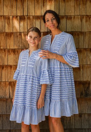 Frau und Mädchen stehen an einer Wand aus Holzschindeln und tragen passende blau-weiß gemusterte knielange Kleider mit Rüschen an Ärmeln und Saum.
