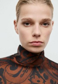 Cuello de tortuga marrón con un diseño ornamentado negro. Textura suave, cuello ajustado y maquillaje minimalista en el rostro del modelo.