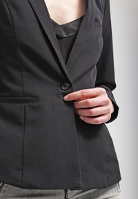 Osoba zapínající knoflík černého blejzru na černém topu s světle šedými kalhotami, viděná od hrudníku po boky na neutrálním pozadí.