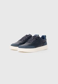 Ein Paar marineblaue Low-Top-Sneakers mit weißen Sohlen, schwarzen Schnürsenkeln und einer grünen Schlaufe auf der Rückseite vor einem schlichten, hellgrauen Hintergrund.