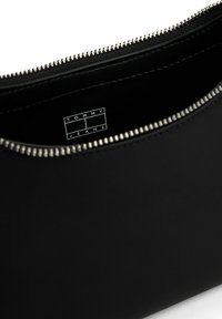 Czarna kopertówka o gładkiej fakturze, z zamkiem błyskawicznym i srebrnymi elementami metalowymi. Wnętrze ozdobione etykietą z napisem "TOMMY JEANS" w prostokątnym wzorze.