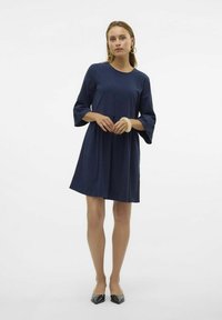 Mörkblå klänning med trekvarts ärmar, lös passform, rundad halsringning och midjesöm. Modellen bär svarta spetsiga skor och armband.