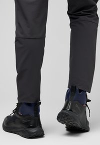Schwarze Wanderschuhe mit einer strukturierten Sohle und blauen Akzenten, kombiniert mit eng anliegenden, dunkelgrauen Hosen. Die Stiefel haben ein hohes Knöbel-Design und sind aus robusten Materialien gefertigt.