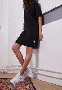 Czarna, oversize sukienka T-shirt z krótkimi rękawami, z bocznym rozcięciem i małą metką marki. W połączeniu z białymi sneakersami na wzorzystym dywaniku.