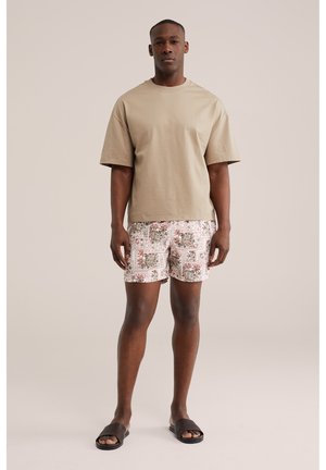 Hombre de pie con camiseta beige de gran tamaño, pantalones cortos blancos con diseños marrones y sandalias negras tipo chancla, sobre fondo liso.