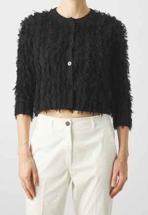 Woman wearing a cropped black textured cardigan with large buttons and white high-waisted pants against a plain background.