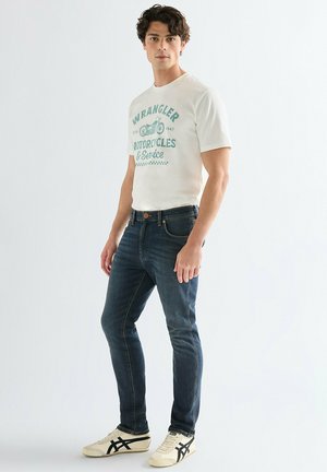 Ung mand stående, iført en hvid t-shirt med grafik og teksten "Wrangler Motorcycles & Service", mørke slim jeans og hvide sneakers.