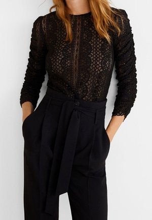 Kobieta nosząca czarną koronkową bluzkę z długim rękawem, włożoną do czarnych spodni z wysokim stanem, z tkaninowym paskiem zawiązanym w talii.
