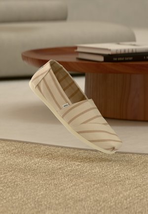 Zapato tipo deportivo sin cordones de lona beige con rayas, colocado sobre una alfombra beige texturizada, mesa de café redonda de madera y libros desenfocados al fondo.