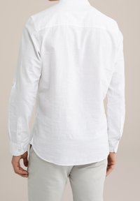 Man som bär en långärmad vit linne skjorta och ljusgrå byxor, sedd bakifrån mot en enfärgad bakgrund.