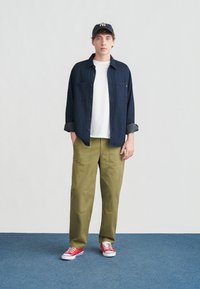 Νέος άνδρας που στέκεται σε μπλε χαλί φοράει μπλε πουκάμισο, λευκό μπλουζάκι, ελιά παντελόνια, κόκκινα αθλητικά παπούτσια και σκούρο καπέλο μπέιζμπολ με λευκό λογότυπο.