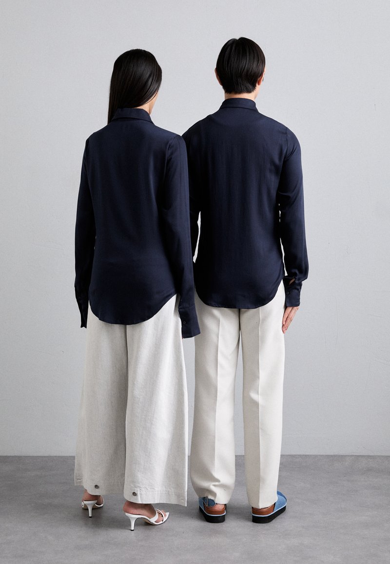 Navy button-up shirts with long sleeves, paired with light beige wide-leg trousers. A detail of white heels and blue footwear is visible.
