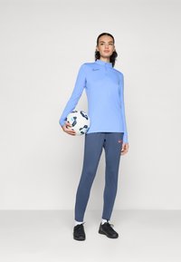 Haut de sport à manches longues bleu clair avec un col haut, associé à un pantalon de sport bleu marine avec un logo Nike rouge. Le modèle tient un ballon de football à motifs.