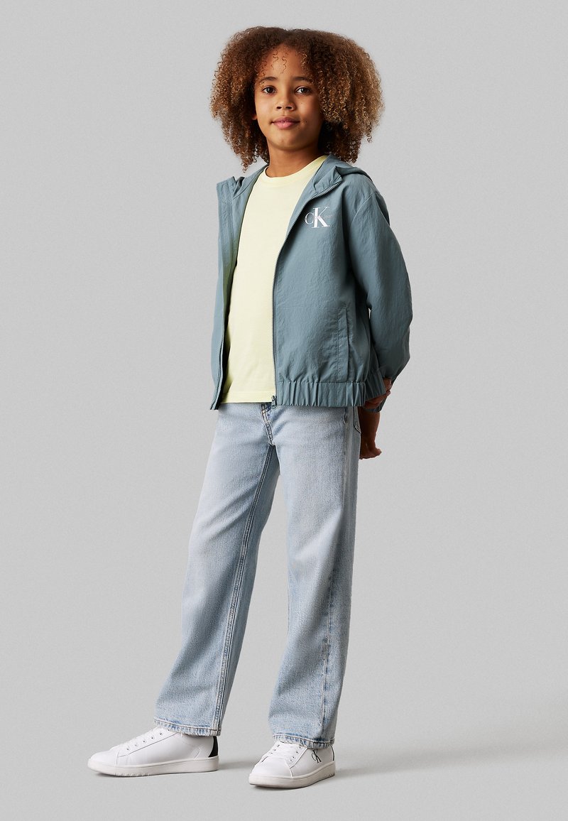Lyseblå zip-jakke med hætte, hvidt logo, parret med en gul t-shirt, lyseblå jeans og hvide sneakers. Glatte teksturer.