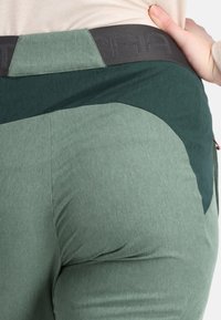 Un pantalon de sport vert présente un tissu texturé avec un accent vert plus foncé et une ceinture noire affichant un logo dans un motif subtil.