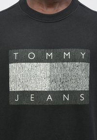 Primer plano de un suéter negro con un logotipo rectangular texturizado que presenta las palabras “TOMMY JEANS” en letras mayúsculas blancas.