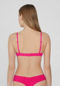 Rosa nahtloser BH mit dünnen verstellbaren Trägern, ausgestattet mit einem Haken- und Ösenverschluss auf der Rückseite. Glatte Stoffstruktur, minimalistisches Design.