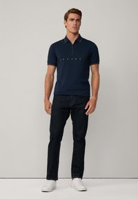Camiseta polo azul marino con texto del logo dorado, mangas cortas y ribete de cuello en contraste, combinada con jeans oscuros y zapatillas blancas.