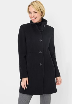 Woman with short blonde hair smiling, wearing a black buttoned coat with high collar and checked pants, standing against a white background.