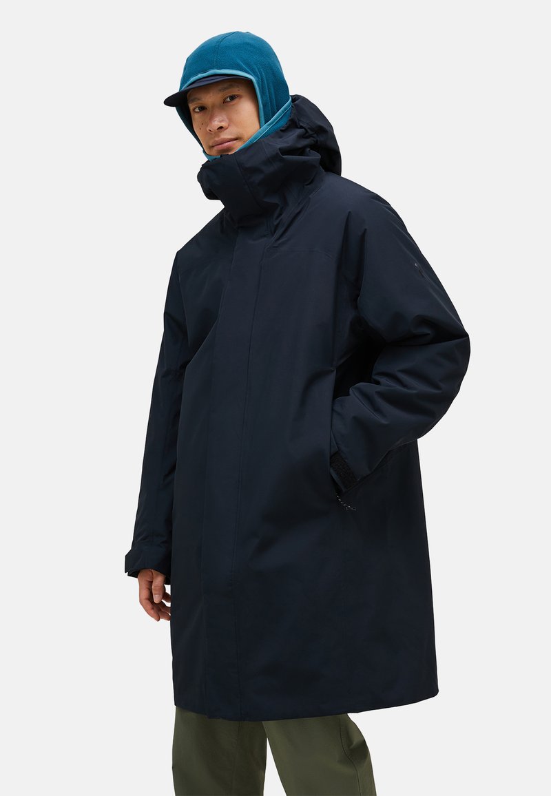 Cappotto impermeabile della marina con colletto alto, zip frontale, tasche laterali e polsini regolabili. Indossato con un berretto teal e cappuccio sotto.