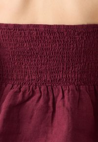 Abito bordeaux senza spalline con texture arricciata sul corpetto e tessuto fluente in vita, caratterizzato da un materiale morbido e leggero.