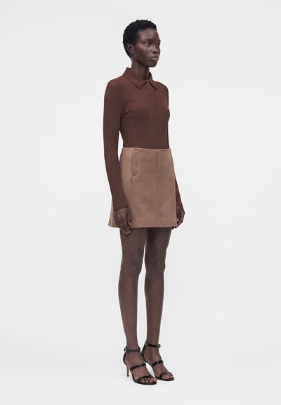 Γυναίκα στέκεται φορώντας καφέ ριμπ μπλουζάκι πόλο με μακριά μανίκια, καμηλό μίνι φούστα από σουέντ με τσέπες και μαύρα σανδάλια με ψηλό τακούνι.