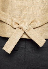 Beige Oberteil aus Stoff mit einem geknoteten Bindedetail hinten. Getragen über dunklen Hosen, zeigt es das strukturierte beige Material.