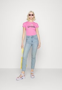 T-shirt rosa ajustada com impressão gráfica, combinada com jeans azul claro. Acessórios incluem uma bolsa de malha amarela e sandálias de plataforma chunky lavanda.