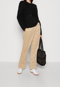 Personne portant un pull noir, un pantalon beige et des baskets blanches, tenant un sac fourre-tout matelassé noir, debout devant un fond uni.