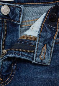 Zipperområdet på denimjeansen med blå denimtyg, kopparorange sömmar, metallknapp och en synlig dragkedja med en mörk tygaccent.