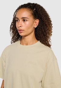 Beige katoenen T-shirt met een ronde hals, korte mouwen en een subtiele geborduurde "HALO"-logo op de borst. Gladde textuur.