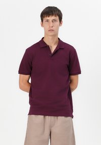 Polo burdeos hecho de algodón, con una tapeta de dos botones y mangas cortas. Combinado con pantalones cortos beige.