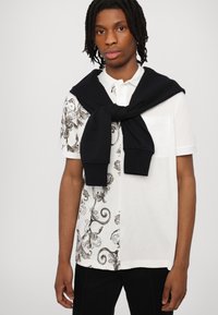 Balta polo krekls ar melnu un pelēku greznu rakstu vienā pusē un vienkāršu priekšējo kabatu; melns džemperis, sasiets ap pleciem.