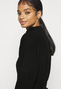 Čierny pletený top s vysokým golierom, s dlhými rukávmi a voľným strihom. Hladká textúra a minimálne dizajnové detaily.